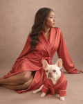 A woman and a small white French Bulldog sit together on a warm beige studio backdrop, both wearing matching satin-like dressing gown robes in a muted brick red with thin light piping. The woman is posed kneeling and looking to the side, while the dog sits in front wearing the same robe style.
