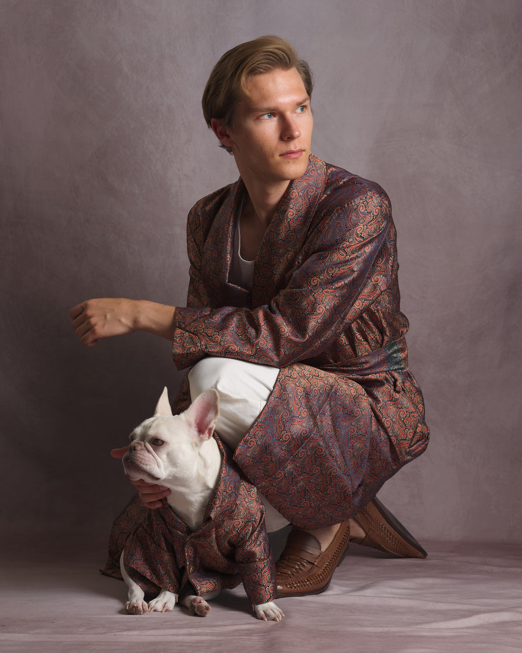 A man crouches in a studio wearing a patterned brown and red kimono dressing gown robe with white pants and brown shoes, while a small white French bulldog sits in front wearing a matching small robe. The background is a solid textured gray studio backdrop.