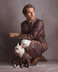 A man crouches in a studio wearing a patterned brown and red kimono dressing gown robe with white pants and brown shoes, while a small white French bulldog sits in front wearing a matching small robe. The background is a solid textured gray studio backdrop.