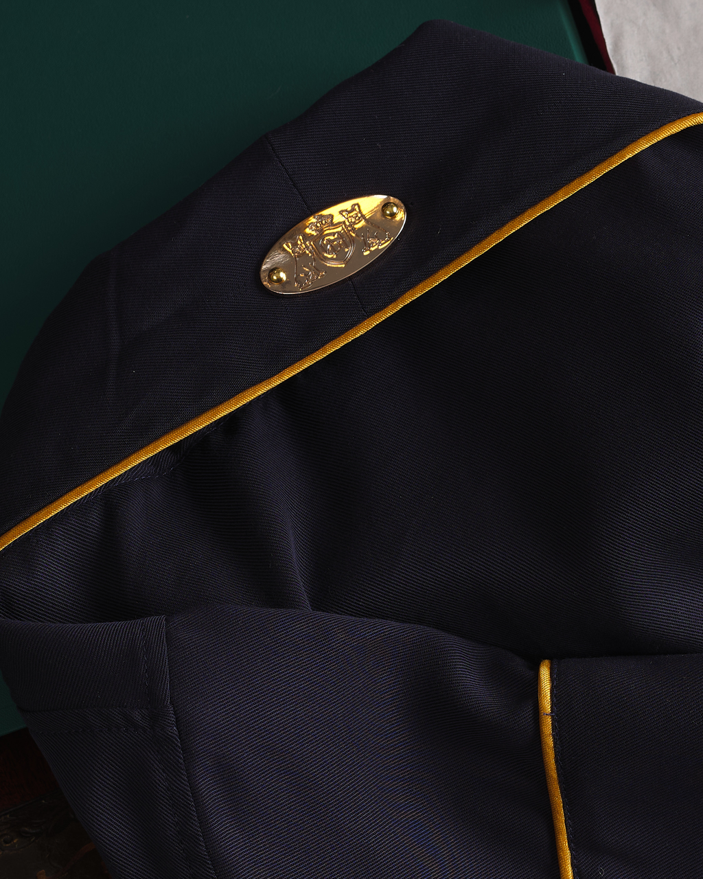 Close-up of a dark navy blue fabric garment with gold piping and a gold oval metal emblem on the shoulder.