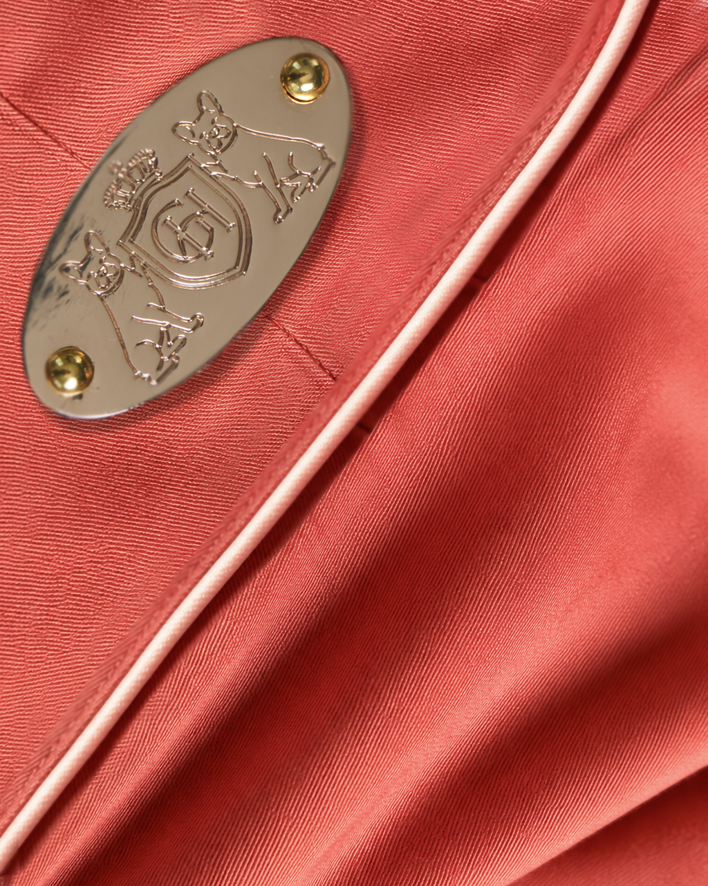 Close-up of a coral pink satin fabric with a light pink piping edge and a silver-tone oval metal emblem secured by gold-tone rivets.
