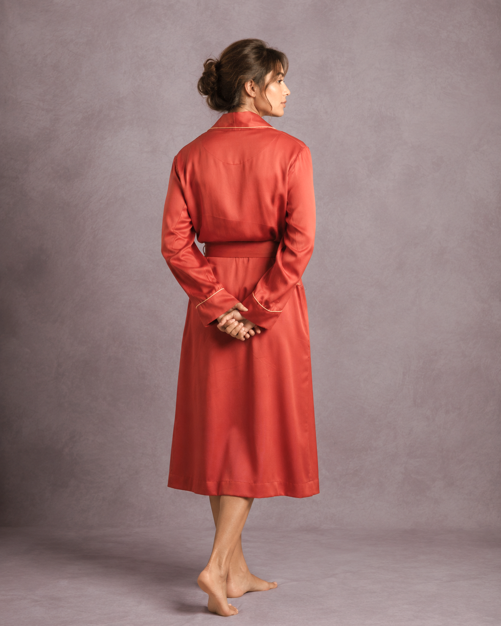 A woman stands barefoot against a textured gray studio backdrop, shown from the back wearing a long-sleeved, belted red dressing gown or robe with a collar and buttoned cuffs.
