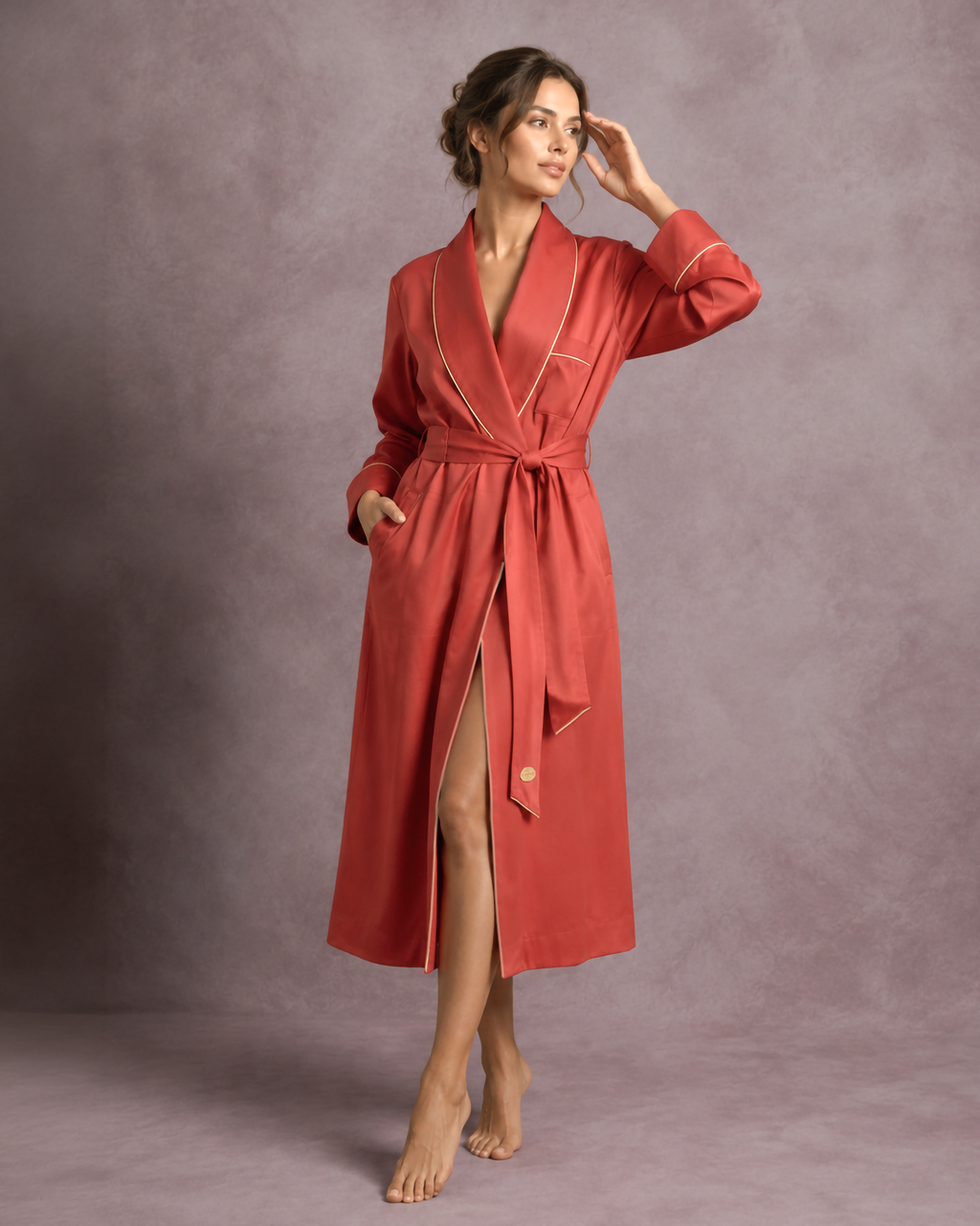 A woman modeling a long terracotta red bathrobe dressing gown  with gold piping and a matching waist tie, standing against a textured mauve studio backdrop.