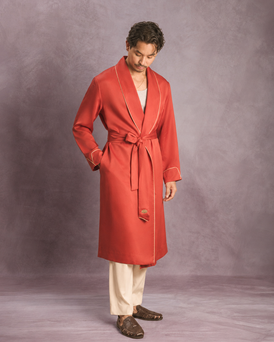 A man wearing a long red robe dressing gown with cream piping and a matching tie belt, paired with cream trousers and brown loafers, standing against a textured purple studio backdrop.