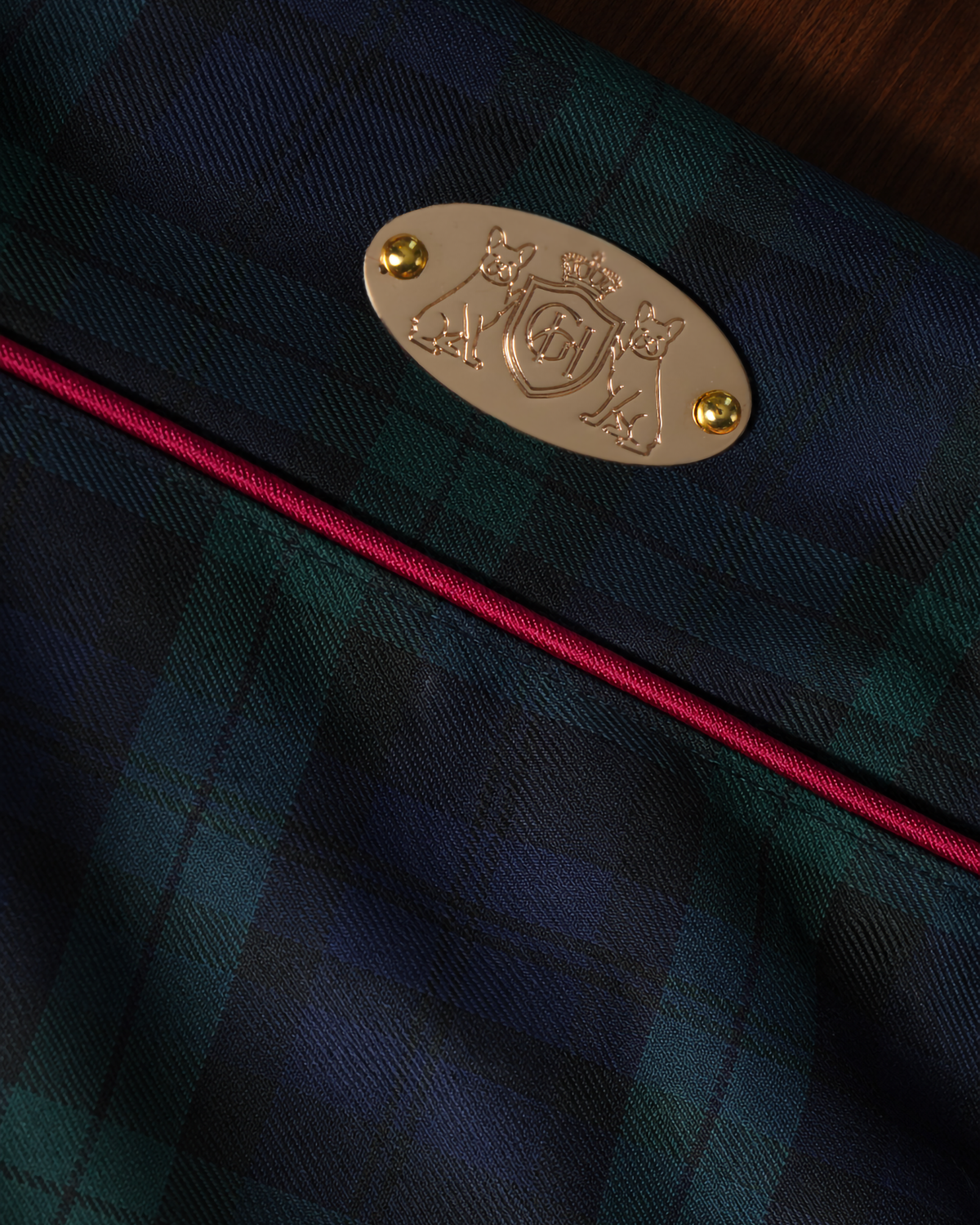 Close-up of a tartan fabric product in dark green and navy plaid with a bright pink piping edge and a gold metal crest badge.