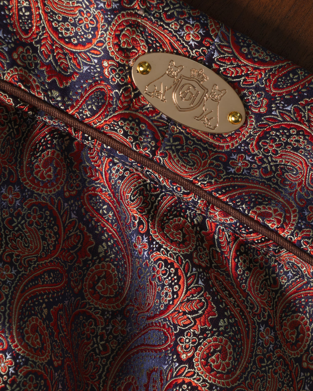 Close-up of a luxurious paisley fabric in navy blue, red, and gold, featuring a polished gold metal crest badge with two gold rivets.