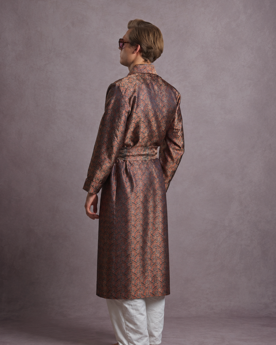 A man wearing a long-sleeved, waist-belted dressing gown in a brown and gold patterned brocade paisley fabric, paired with white trousers, photographed from the back against a textured gray studio backdrop.