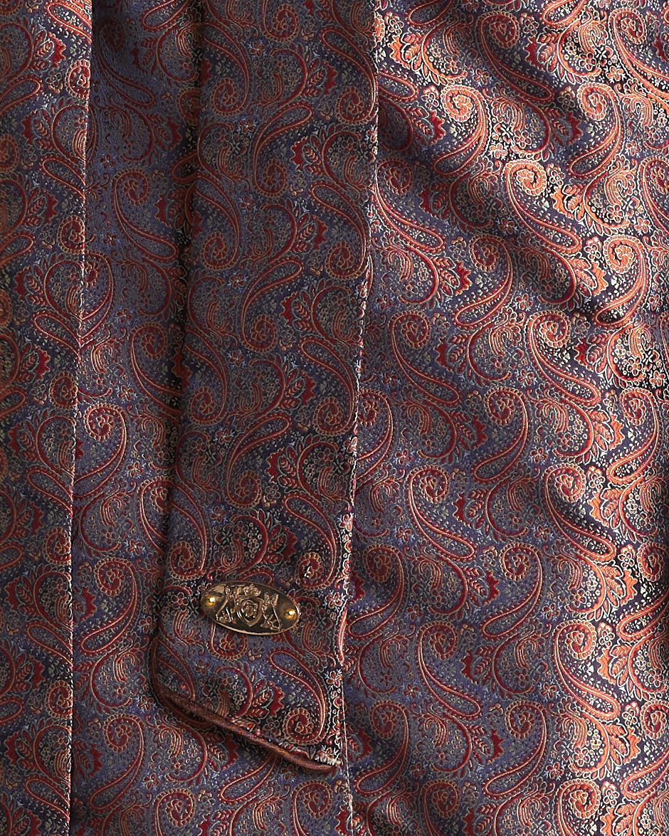Close-up of a paisley patterned fabric garment, likely a shirt or jacket, featuring an intricate paisley design in muted blue, rust red, and gold tones, with a decorative gold button on the chest pocket.