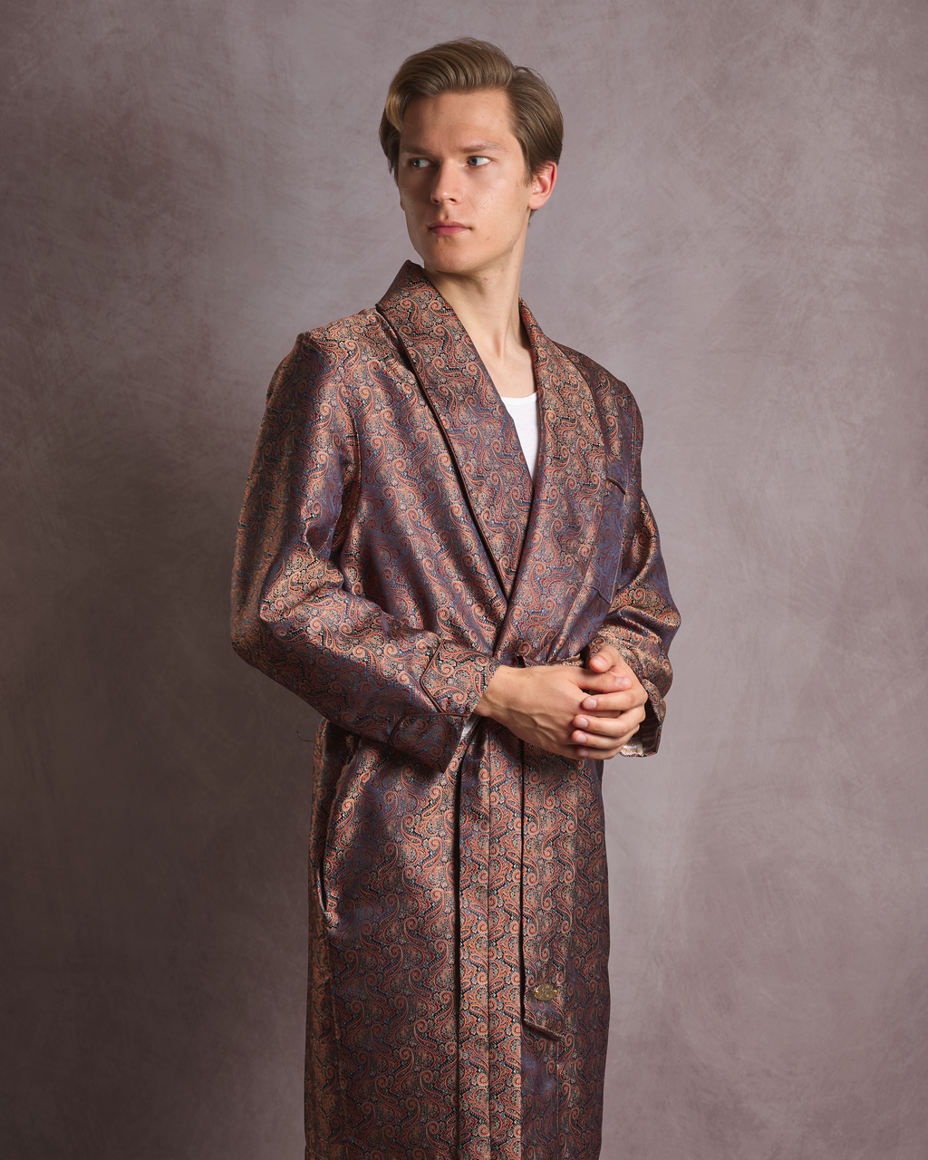 A man wearing a long paisley patterned bathrobe dressing gown with a shawl collar and waist tie, standing against a textured gray studio backdrop.