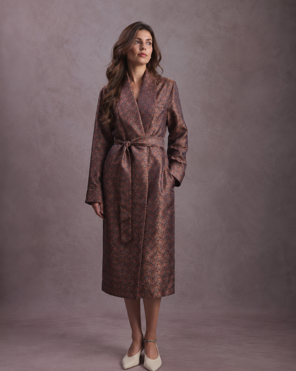 A woman modeling a long brown paisley robe dressing gown with a matching tie belt, standing against a textured mauve studio backdrop.