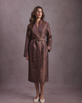 A woman modeling a long brown paisley robe dressing gown with a matching tie belt, standing against a textured mauve studio backdrop.