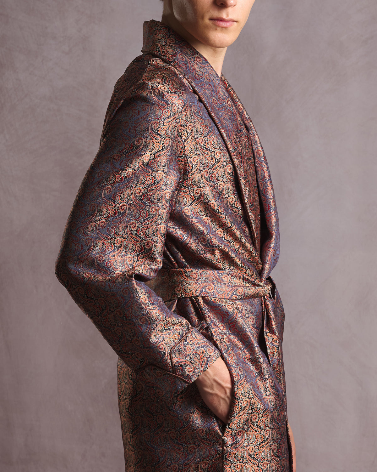 A man wearing a luxurious patterned paisley silk dressing gown robe in brown, gold, and blue, shown from a side profile against a textured gray studio background.